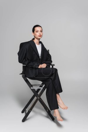 Full length of barefoot woman in striped suit sitting on folding chair on grey background 