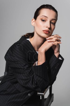 Téléchargez les photos : Portrait de femme brune élégante posant sur chaise isolée sur gris - en image libre de droit