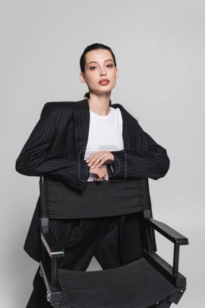 Téléchargez les photos : Jolie femme brune debout près d'une chaise pliante et regardant la caméra isolée sur gris - en image libre de droit