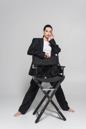 Longitud completa de la mujer de moda en traje posando cerca de la silla plegable sobre fondo gris 