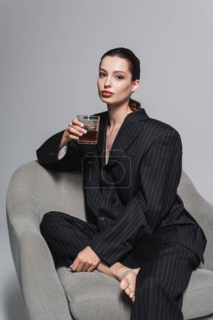 Foto de Elegante modelo en traje negro sosteniendo vaso de whisky mientras está sentado en un sillón sobre fondo gris - Imagen libre de derechos