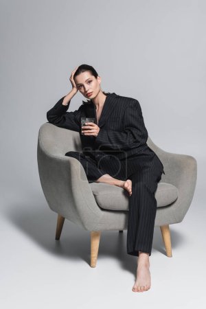 Mujer descalza en traje negro sosteniendo un vaso de whisky mientras está sentada en un sillón sobre fondo gris 