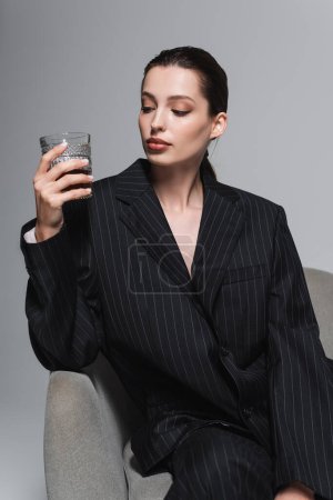 Foto de Mujer con estilo en chaqueta a rayas mirando el vaso de whisky en sillón aislado en gris - Imagen libre de derechos