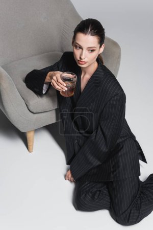 Overhead view of woman in suit holding whiskey in glass near armchair on grey background 