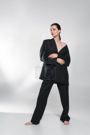 Téléchargez les photos : Pleine longueur de femme en veste noire avec épaule nue regardant loin sur fond gris abstrait - en image libre de droit