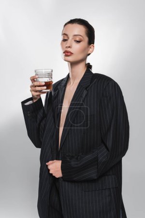 Foto de Mujer joven con estilo en chaqueta a rayas con whisky sobre fondo gris abstracto - Imagen libre de derechos