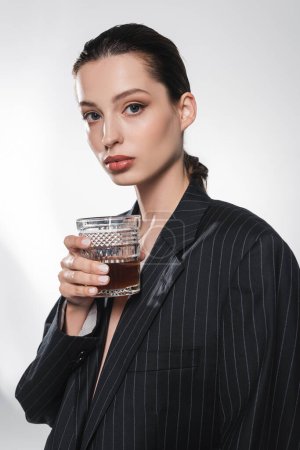 Foto de Mujer joven de moda con chaqueta negra sosteniendo un vaso de whisky y mirando a la cámara sobre fondo gris - Imagen libre de derechos