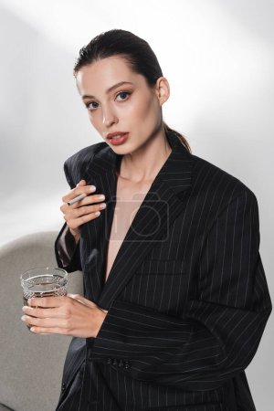 Femme élégante en veste noire tenant un verre de whisky et de cigarette près du fauteuil sur fond gris 