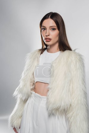 Stylish woman in white faux fur jacket looking at camera on grey background 