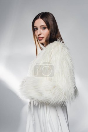 Photo for Stylish brunette woman in faux fur jacket looking at camera on abstract grey background - Royalty Free Image