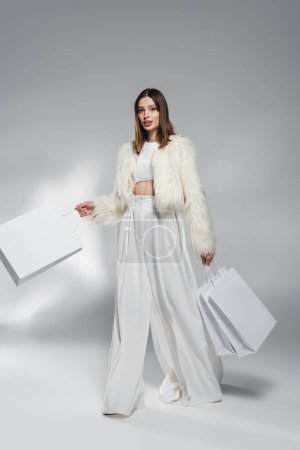 Fashionable woman in faux fur jacket and pants holding shopping bags on abstract grey background 