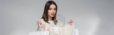 Stylish model in white faux fur jacket holding shopping bags on grey background, banner 