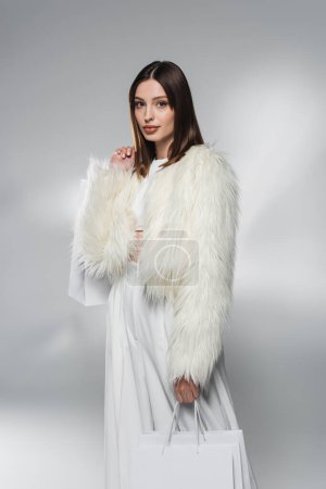 Trendy brunette woman in white faux fur jacket holding shopping bags on abstract grey background 