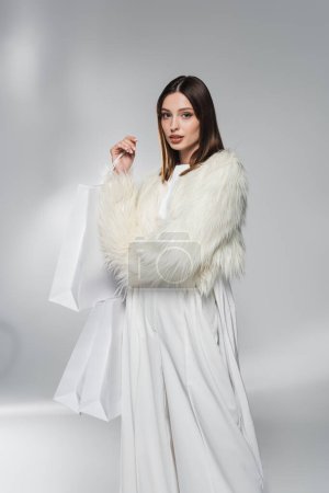 Photo for Fashionable woman in white faux fur jacket holding shopping bags and looking at camera on abstract grey background - Royalty Free Image