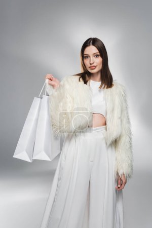 Fashionable woman in faux fur jacket holding white shopping bags on abstract grey background 