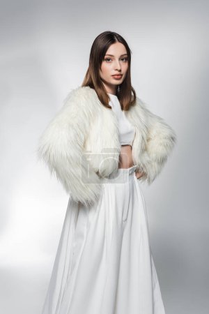 Téléchargez les photos : Femme tendance en vêtements blancs et veste en fausse fourrure tenant la main sur les hanches sur fond gris abstrait - en image libre de droit