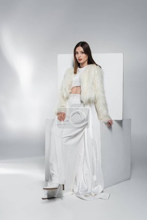 Photo for Stylish young woman in white clothes and faux fur jacket posing near cubes on abstract grey background - Royalty Free Image