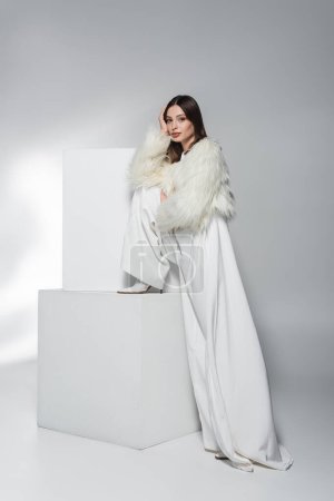 Stylish woman in white clothes and faux fur jacket looking at camera near cubes on abstract grey background 