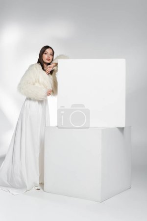 Stylish woman in white faux fur jacket looking at camera near cubes on abstract grey background 