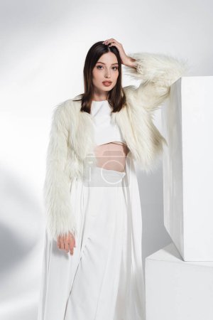 Photo for Pretty young woman in white faux fur jacket and crop top leaning on cube while posing on grey - Royalty Free Image