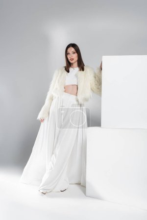 Photo for Full length of young woman in faux fur jacket and total white outfit leaning on cubes while posing on grey - Royalty Free Image
