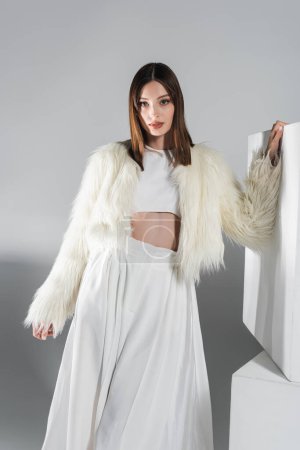 Photo for Young woman in faux fur jacket and total white outfit posing near cubes on grey - Royalty Free Image