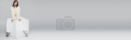 Téléchargez les photos : Pleine longueur de femme élégante en fausse veste de fourrure et tenue blanche totale assise sur cube sur gris, bannière - en image libre de droit