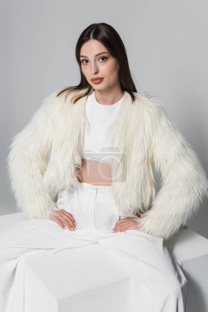 young woman in trendy faux fur jacket and total white outfit sitting with hands on hips on cube isolated on grey