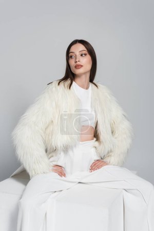stylish woman in white faux fur jacket sitting with hands on hips on cube isolated on grey