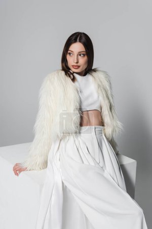 Téléchargez les photos : Jeune femme à la mode veste en fausse fourrure et tenue blanche totale appuyée sur cube sur gris - en image libre de droit