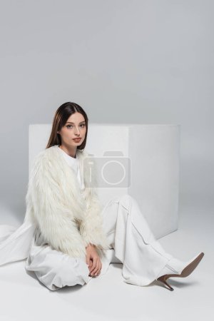 Photo for Full length of trendy woman in faux fur jacket and total white outfit sitting near cube on grey - Royalty Free Image