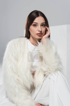Photo for Young woman in white faux fur jacket looking at camera near cube isolated on grey - Royalty Free Image