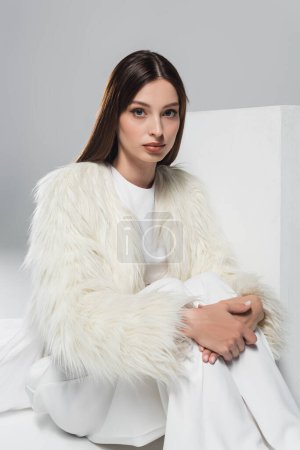 Photo for Stylish young woman in white faux fur jacket looking at camera near cube on grey - Royalty Free Image