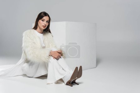 Foto de Longitud completa de la mujer joven con estilo en chaqueta de piel sintética blanca mirando a la cámara cerca del cubo sobre fondo gris - Imagen libre de derechos