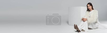 Téléchargez les photos : Pleine longueur de jeune femme élégante en fausse fourrure blanche veste regardant la caméra près du cube sur gris, bannière - en image libre de droit