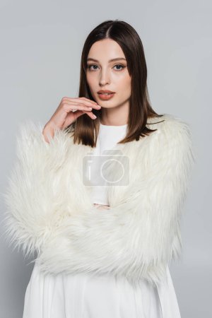 Téléchargez les photos : Portrait de jolie jeune femme en veste blanche élégante en fausse fourrure regardant la caméra isolée sur gris - en image libre de droit
