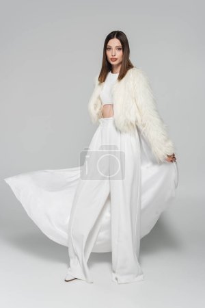 Foto de Longitud completa de la mujer de moda en traje totalmente blanco y chaqueta de piel sintética de pie en gris - Imagen libre de derechos