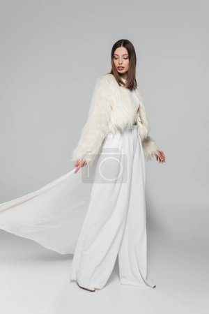 full length of pretty woman in totally white outfit and faux fur jacket posing on grey