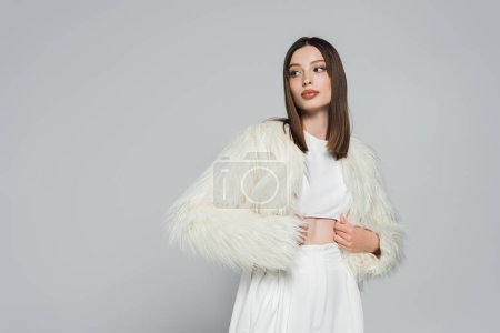 young woman in totally white outfit and trendy faux fur jacket posing isolated on grey