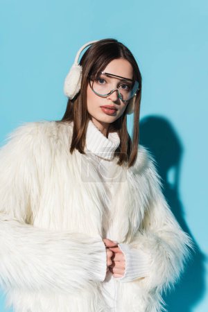 Photo for Trendy woman in earmuffs and eyeglasses posing in faux fur jacket while looking at camera on blue - Royalty Free Image