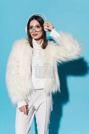 stylish young woman in trendy eyeglasses and faux fur jacket adjusting winter earmuffs on blue 
