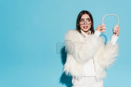 amazed young woman in eyeglasses and faux fur jacket holding winter earmuffs on blue 