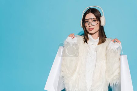 young woman in earmuffs and stylish faux fur jacket holding shopping bags on blue background 