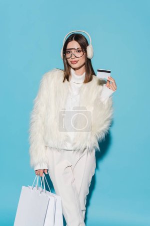 pretty woman in winter earmuffs and stylish faux fur jacket holding credit card and shopping bags on blue 
