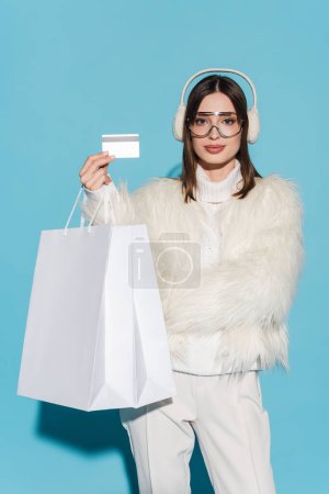 Téléchargez les photos : Jeune femme en lunettes et veste élégante en fausse fourrure tenant carte de crédit et sacs à provisions sur bleu - en image libre de droit
