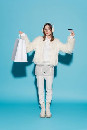 full length of surprised woman in eyeglasses and stylish faux fur jacket holding credit card and shopping bags on blue