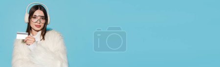Téléchargez les photos : Jeune femme heureuse en lunettes et veste élégante en fausse fourrure tenant la carte de crédit isolée sur bleu, bannière - en image libre de droit