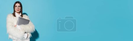 Téléchargez les photos : Femme élégante dans des lunettes de vue à la mode et veste élégante en fausse fourrure tenant ordinateur portable sur bleu, bannière - en image libre de droit