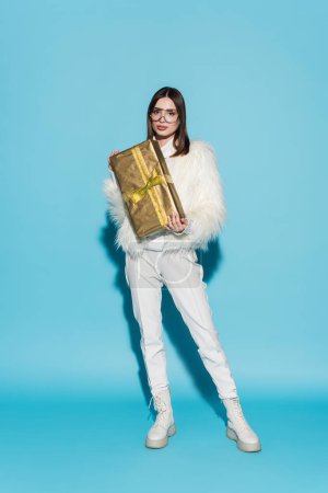 full length of stylish woman in faux fur jacket and eyeglasses holding christmas present on blue