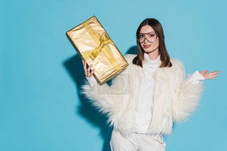stylish woman in faux fur jacket and eyeglasses holding christmas present on blue background